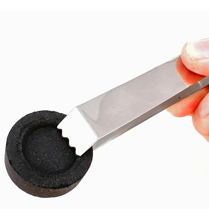 Hand mit Metallzange beim Greifen einer runden schwarzen Kohletablette